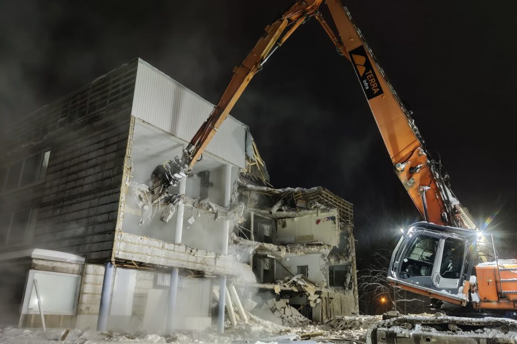 Rakennusta puretaa pimeässä työvalojen loisteessa. Oranssi työkone. 